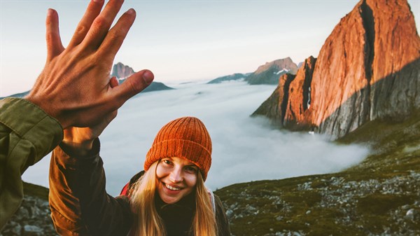 Ženska z oranžno kapo se smeji in daje petko ob razgledu na gore nad morjem megle.