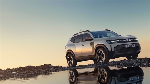 Na sliki je nova Dacia Duster v svetli barvi, parkirana na skalnatem terenu ob vodi, v kateri se odseva vozilo.