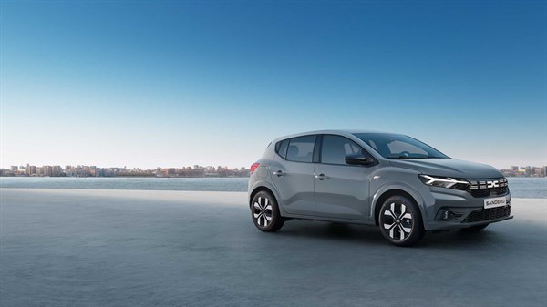 Na sliki je nova Dacia Sandero v sivi barvi, parkirana ob obali z mestnim ozadjem in modrim nebom. 