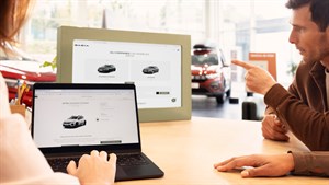 Na sliki sta dve osebi, ki v Dacia salonu skupaj izbirata vozilo prek konfiguratorja na računalniku in zaslonu. V ozadju je razstavljen rdeč Dacia model. 
