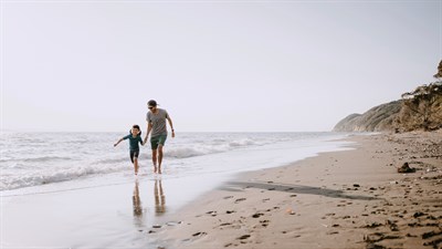 Oče in otrok hodita po mokrem delu peščene plaže ob morju.