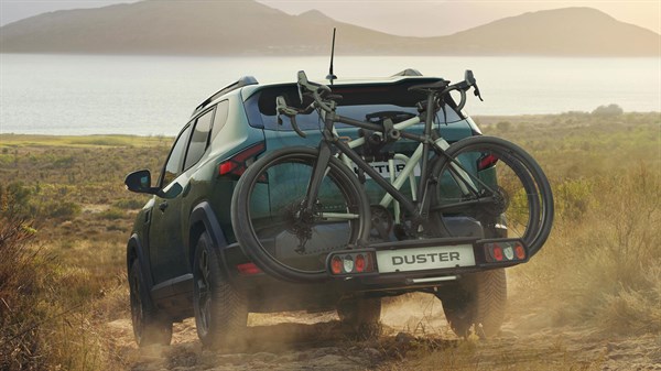 Dacia Duster temno zelene barve, zadnji pogled na makadamski cesti, s kolesom na nosilcu za kolesa na zadnjih vratih.
