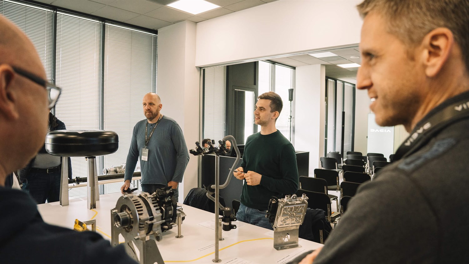 Skupina ljudi si ogleduje avtomobilske komponente na delavnici v notranjem prostoru.