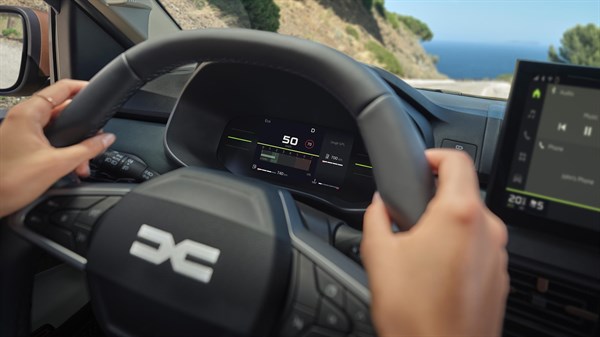 Voznik drži volan vozila Dacia z digitalnim prikazovalnikom pri vožnji ob morju.
