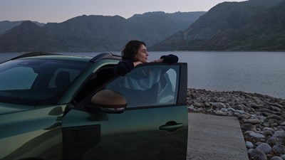 Ženska naslonjena na odprta vrata zelenega Dacia Duster ob jezeru z gorskim ozadjem.