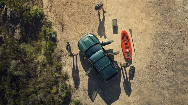 Zračni pogled na temno zeleni Dacia Duster parkiran na makadamski površini z odprtimi vrati, ob njem so ljudje, prtljaga in rdeč kajak.
