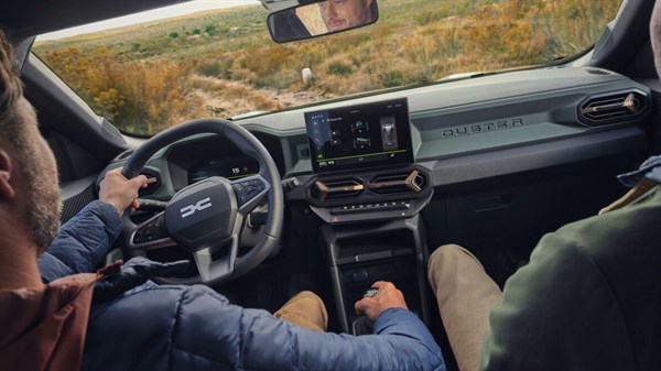 Pogled iz notranjosti Dacia Duster med vožnjo po neutrjeni poti, voznik drži volan in prestavno ročico, na zaslonu prikaz vožnje po terenu.