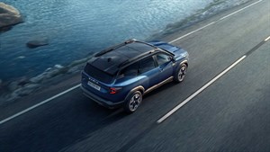 Moder SUV vozila Dacia Bigster vozi po obalni cesti ob vodi. Vozilo je prikazano iz ptičje perspektive, v gibanju.