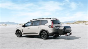 Na sliki je svetel Dacia Jogger, prikazan v bočnem zadnjem pogledu, parkiran ali ustavljen na svetli peščeni podlagi z nežnim naravnim ozadjem.