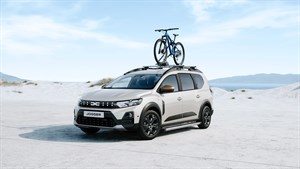 Na sliki je svetel Dacia Jogger, prikazan v bočnem prednjem pogledu, parkiran ali ustavljen na svetli peščeni podlagi z nežnim naravnim ozadjem ter kolesom na strešnih nosilcih.
