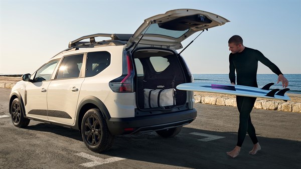 Sivi Dacia Jogger z odprtim prtljažnikom in deskarjem ob morju.