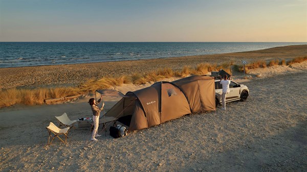 Beli Dacia Jogger s šotorom pritrjenim na prtljažna vrata na plaži ob sončnem zahodu.