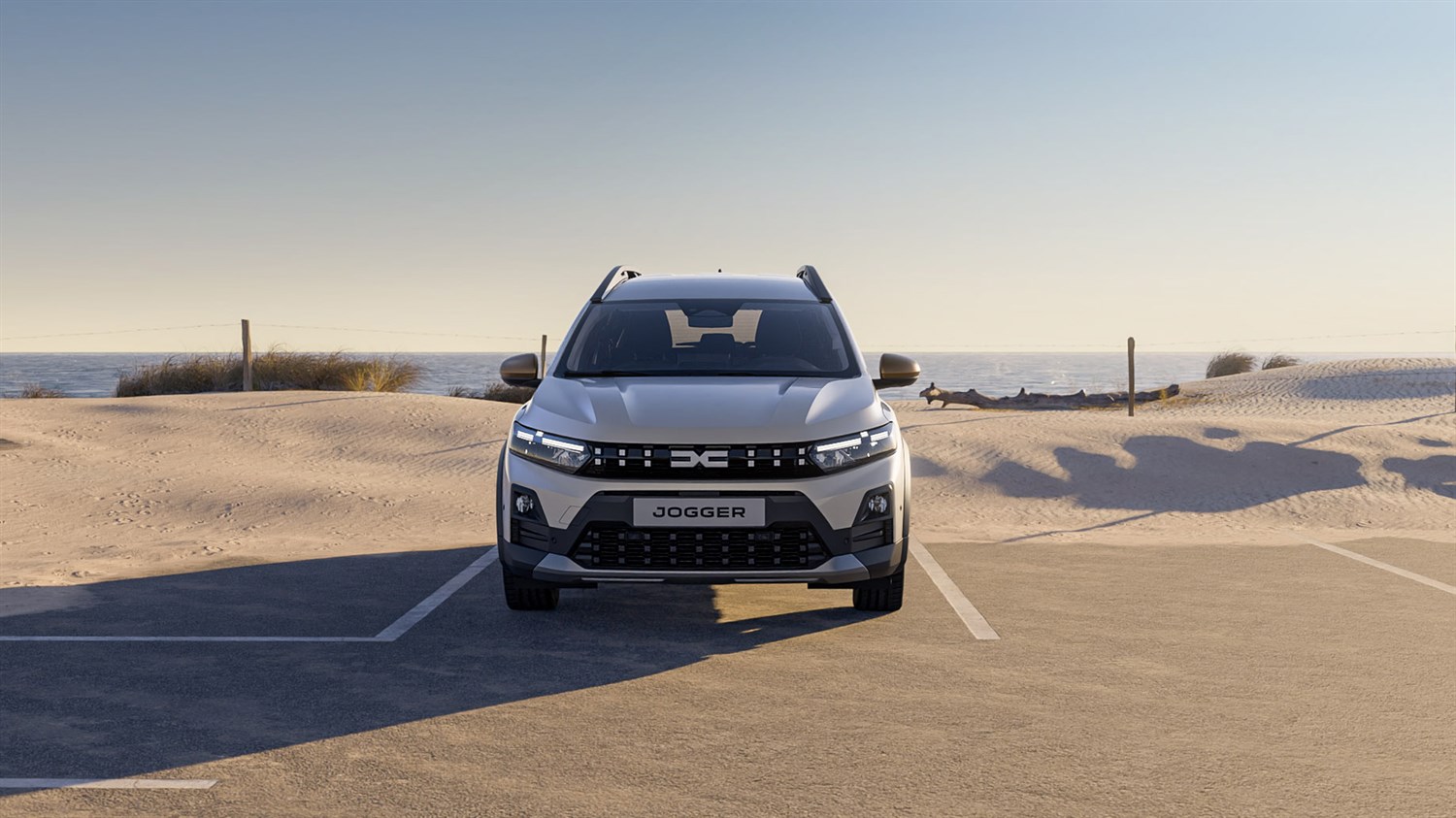 Na sliki je sprednji pogled na Dacio Jogger, parkirano na urejenem betonskem parkirišču ob morski obali. Ozadje tvorijo peščene sipine, nizko rastlinje in mirna morska gladina pod jasnim nebom.