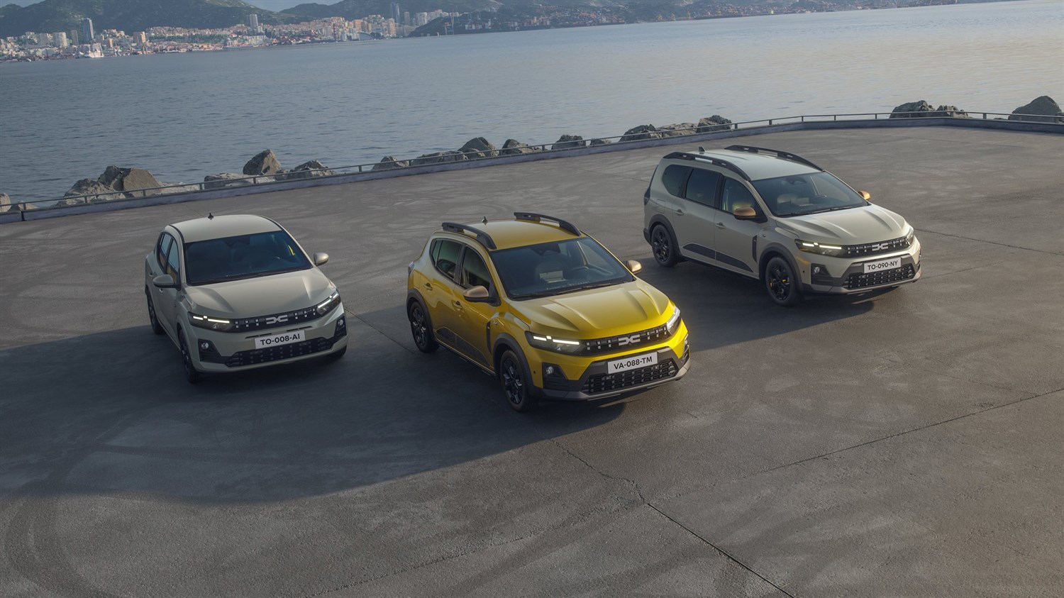 Trije modeli vozil Dacia, siv Sandero, rumen Sandero Stepway, siv Jogger - parkirani ob obali z razgledom na morje in mesto v ozadju.