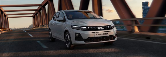 Na sliki je bel Dacia Sandero, ki vozi po mostu v času sončnega zahoda.