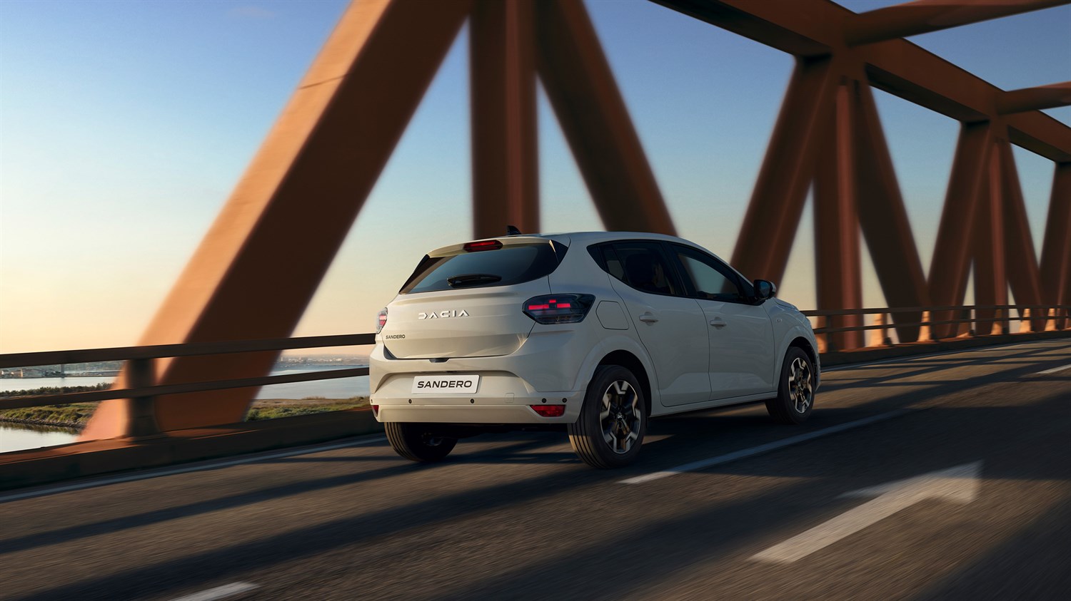 Na sliki je bel Dacia Sandero, ki vozi po mostu v času sončnega zahoda.