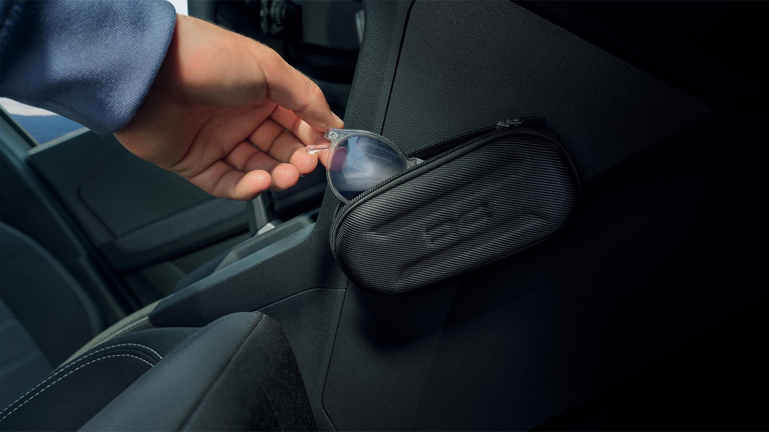 Na sliki je notranjost vozila s poudarkom na majhni črni torbici, ki je pritrjena ob sedežu. Torbica je odprta in iz nje štrlijo sončna očala z prozornim okvirjem, ki jih oseba ravno jemlje ali odloži.