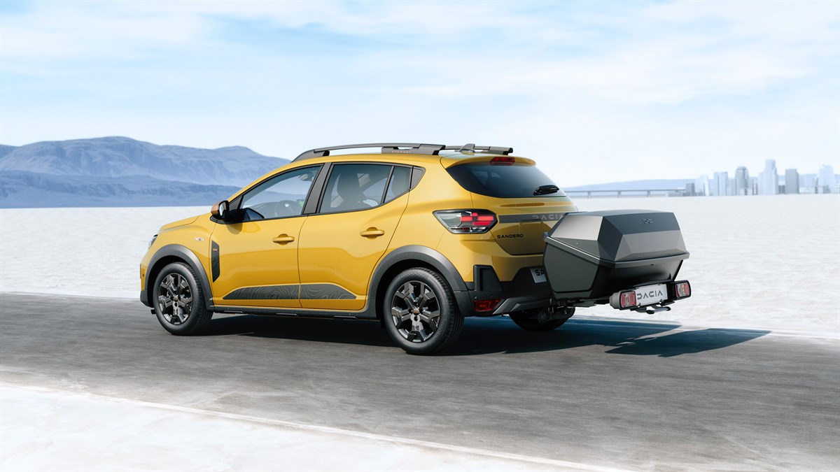 Na sliki je rumen Dacia Sandero Stepway, prikazan v vožnji po ravni, svetli cesti v odprti pokrajini. Vozilo ima nameščen priklopni kovček na vlečni kljuki, ki služi kot dodatni prostor za prtljago.