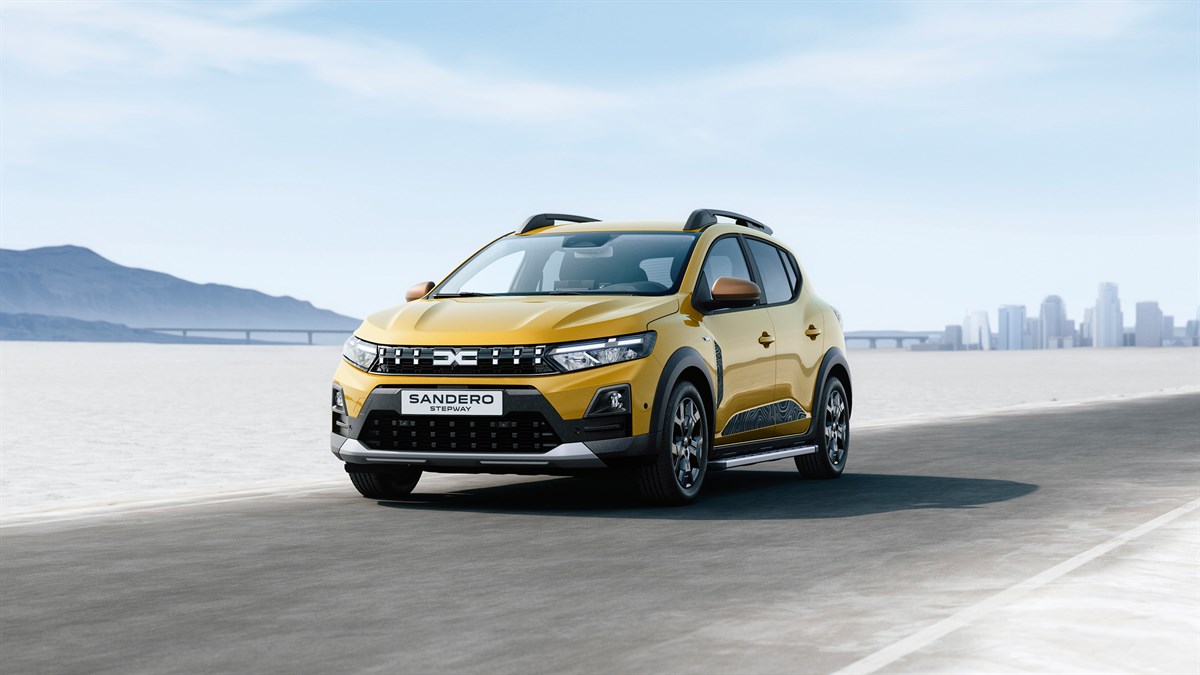 Na sliki je rumen Dacia Sandero Stepway v vožnji po ravni, svetli cesti sredi odprte pokrajine. Vozilo je obrnjeno rahlo proti desni in poudarja robusten videz s črnimi obrobami, strešnimi nosilci in zaščitnimi elementi na odbijaču.