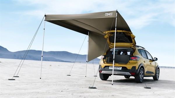 Na sliki je rumen Dacia Sandero Stepway, parkiran na svetlem, peščenem območju. Prtljažna vrata so dvignjena, iz zadnjega dela pa je nameščena velika zunanja tenda, ki je pritrjena na strešne nosilce vozila.
