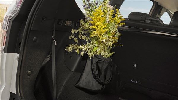 Na sliki je prtljažni prostor vozila, kjer je na levi strani nameščena majhna črna viseča torbica. V njej je šopek cvetja z rumenimi in vijoličnimi cvetovi ter zelenjem