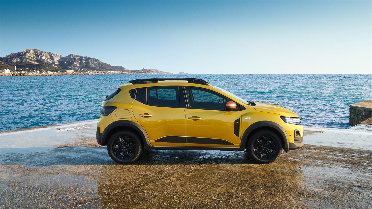 Na sliki je rumeni Dacia Sandero Stepway, parkiran ob morski obali ob sončnem zahodu. Avto stoji na mokri betonski površini, vidno opazovalcu z boka.