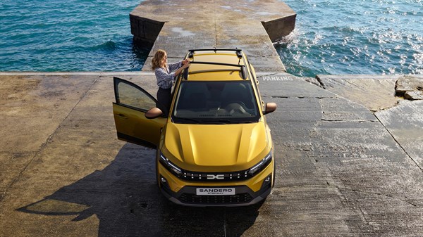 Na sliki je rumena Dacia Sandero Stepway, parkirana ob robu pomola tik nad morsko gladino. Vozilo je fotografirano od zgoraj, kar jasno pokaže strešne nosilce, streho in sprednji del.
