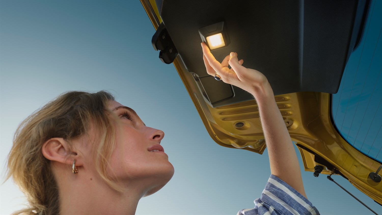 Na sliki je oseba, ki stoji pod odprtimi zadnjimi vrati vozila in z roko sega proti notranji lučki, nameščeni na spodnji strani prtljažnih vrat.