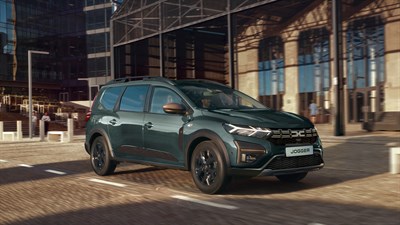 Dacia Jogger v temno zeleni barvi vozi po tlakovanem mestnem cestišču mimo sodobne zastekljene stavbe.