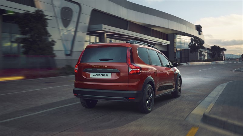 Dacia Jogger v rdeči barvi med vožnjo po mestni ulici mimo sodobne arhitekturne stavbe, pogled od zadaj.