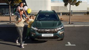Dacia Jogger v temno zeleni barvi stoji na parkirišču, pred njim hodi nasmejan moški z dvema otrokoma, eden sedi na njegovih ramenih, drugi drži balone.