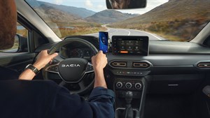 Pogled iz notranjosti vozila Dacia Jogger med vožnjo po podeželski cesti, voznik drži volan in pametni telefon, na sredinski konzoli je prikazan zaslon z navigacijo.