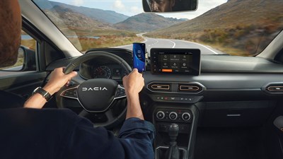 Pogled iz notranjosti med vožnjo po podeželski cesti, voznik drži volan in pametni telefon, na sredinski konzoli je prikazan zaslon z navigacijo.