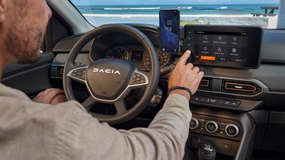 Voznik v Dacia Sandero Stepway upravlja multimedijski zaslon na dotik, ob njem je pametni telefon nameščen v držalu.